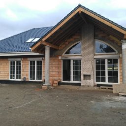 PHU SEBMARK Marek Olejnik - Budynek w trakcie budowy z widocznymi oknami z białymi ramami, ceglaną elewacją, dachem z ciemnej dachówki z oknami dachowymi oraz kolumnami przed wejściem, na pierwszym planie ziemia.