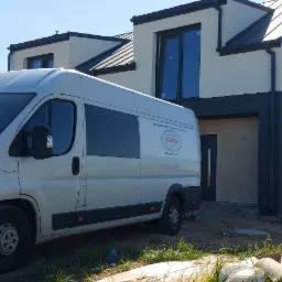 Nowoczesny dom w trakcie wykończenia elewacji, z zaparkowanym obok samochodem dostawczym firmy budowlanej z widocznym logo i danymi kontaktowymi na boku pojazdu.