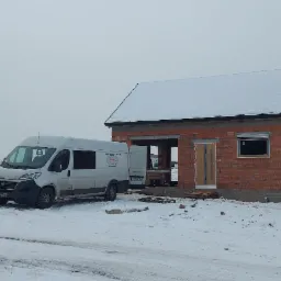 W zimowej scenerii w Kaliszu, budynek w trakcie budowy z zamontowanymi oknami z roletami zewnętrznymi oraz częściowo zamontowanymi drzwiami wejściowymi. Na pierwszym planie zaparkowane busy dostawcze.