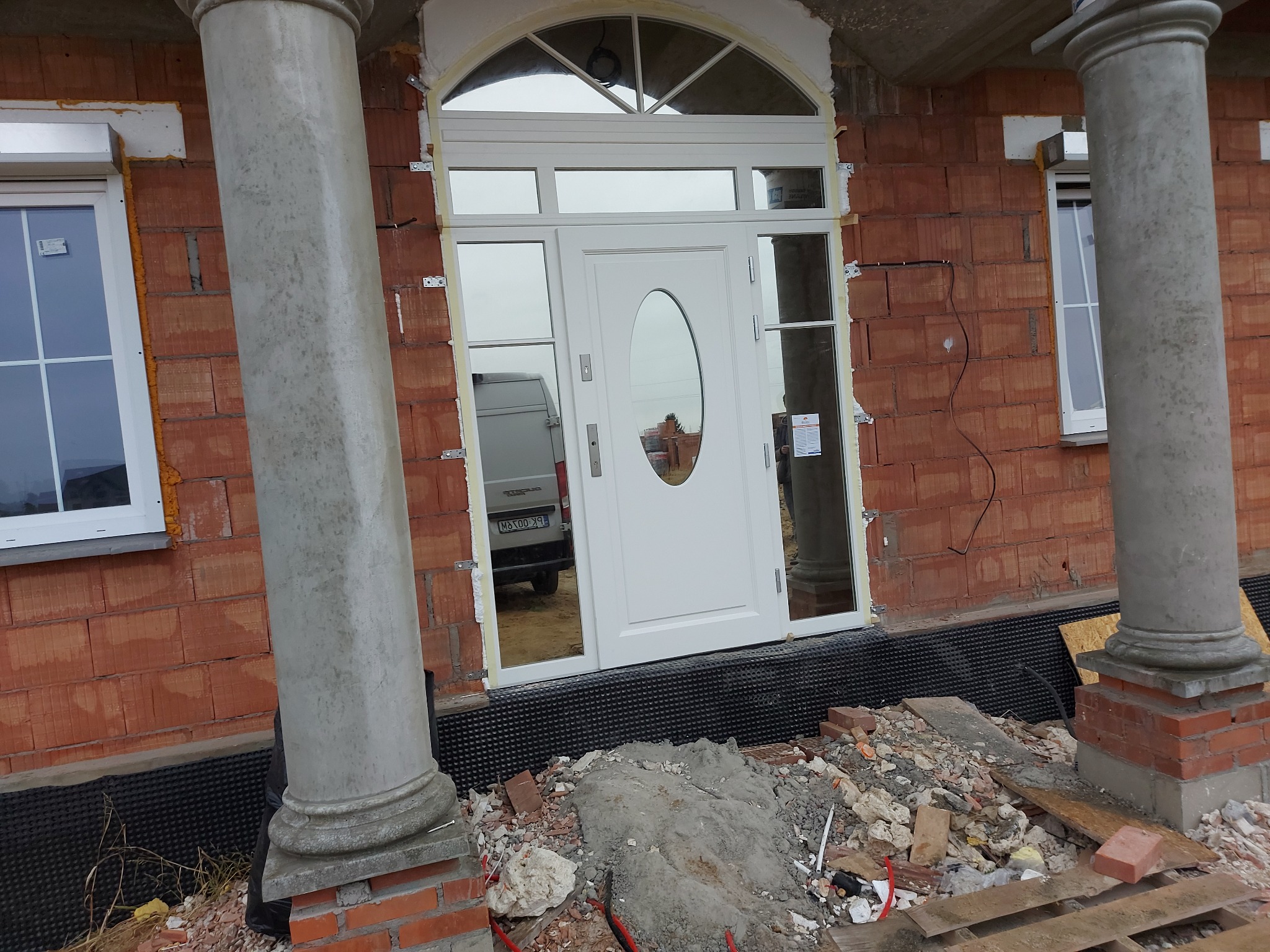 Białe drzwi wejściowe z owalnym przeszkleniem, zamontowane w surowym stanie budynku z cegły, widoczne kolumny i okna, w tle zaparkowany samochód dostawczy.