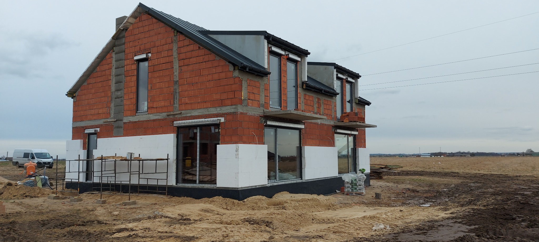 Surowy stan domu jednorodzinnego w trakcie ocieplania ścian zewnętrznych styropianem, widoczne rusztowanie, paleta z materiałami budowlanymi oraz częściowo zamontowane okna z roletami zewnętrznymi.