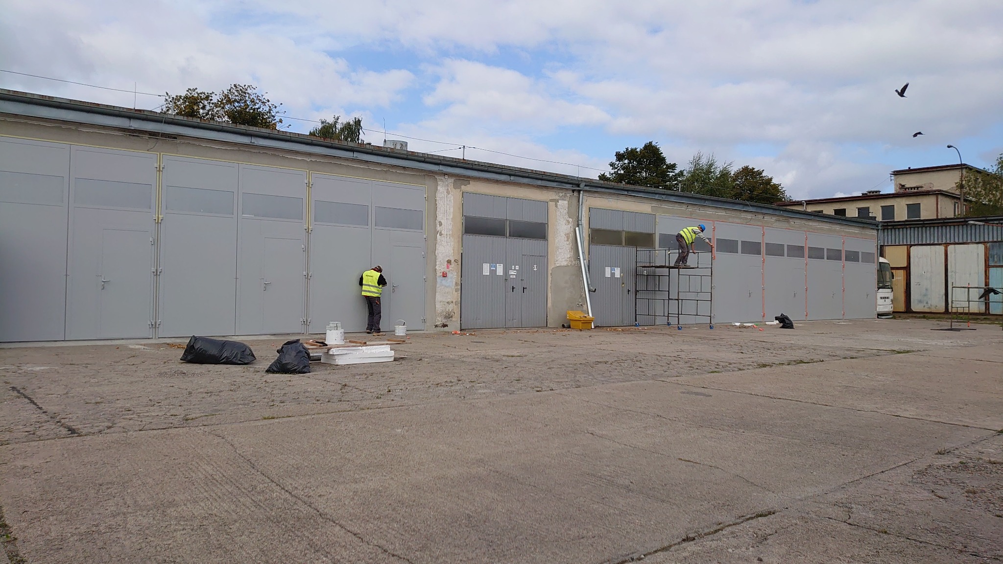 Malowanie szarych drzwi garażowych: Dwóch pracowników w kamizelkach odblaskowych maluje metalowe drzwi garażowe na zewnątrz budynku, jeden z drabiny, drugi z ziemi. Widoczne worki na śmieci, puszki...