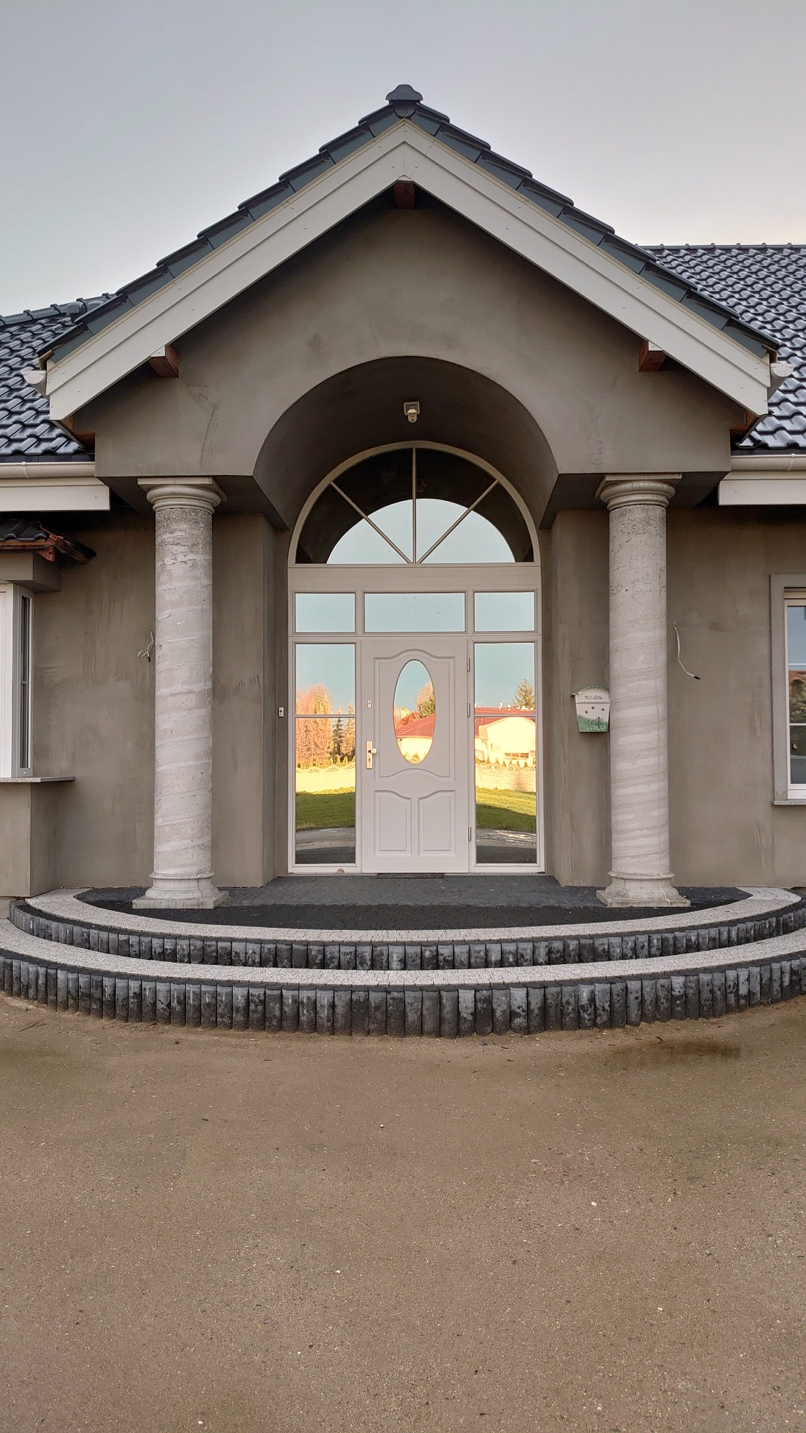 Białe drzwi wejściowe z owalnym przeszkleniem, otoczone kolumnami i łukiem, widoczne z zewnątrz na tle domu z szarą elewacją i schodami z kostki brukowej.