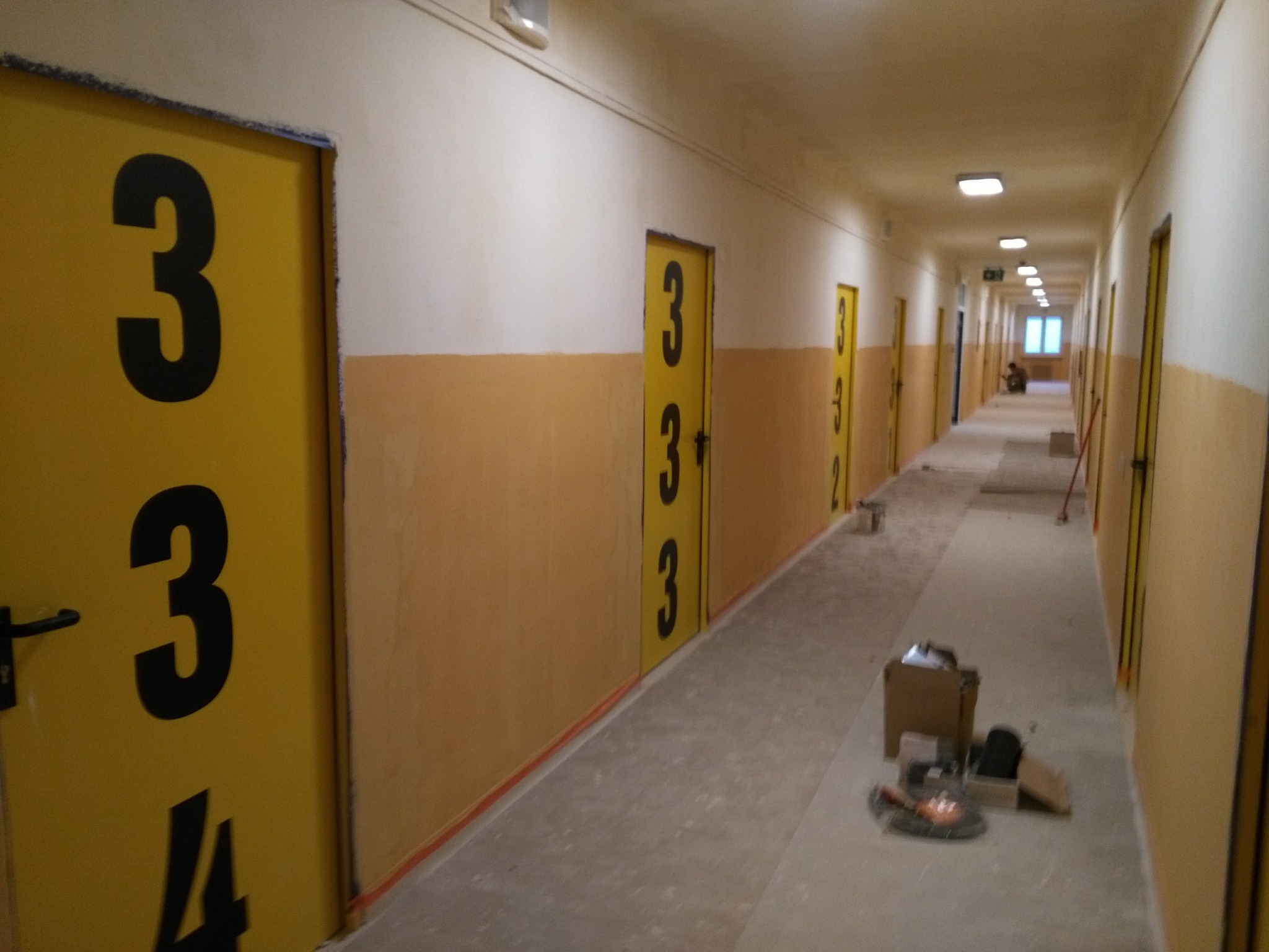 Długi korytarz z żółtymi drzwiami oznaczonymi czarnymi numerami, widoczne ślady remontu, kartony z narzędziami na podłodze, malowanie ścian w dwóch kolorach: biały i żółty.
