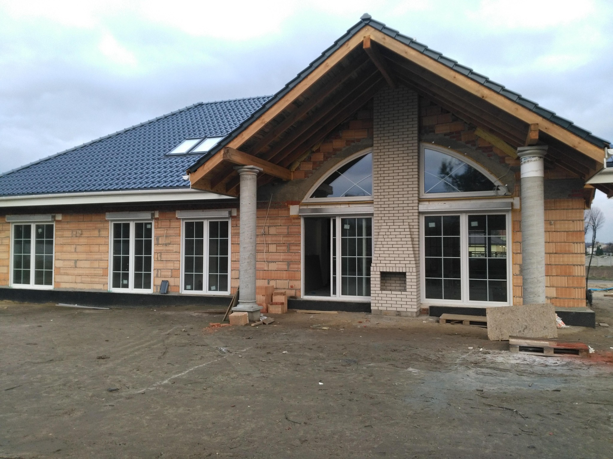 Budynek w trakcie budowy z widocznymi oknami z białymi ramami, ceglaną elewacją, dachem z ciemnej dachówki z oknami dachowymi oraz kolumnami przed wejściem, na pierwszym planie ziemia.