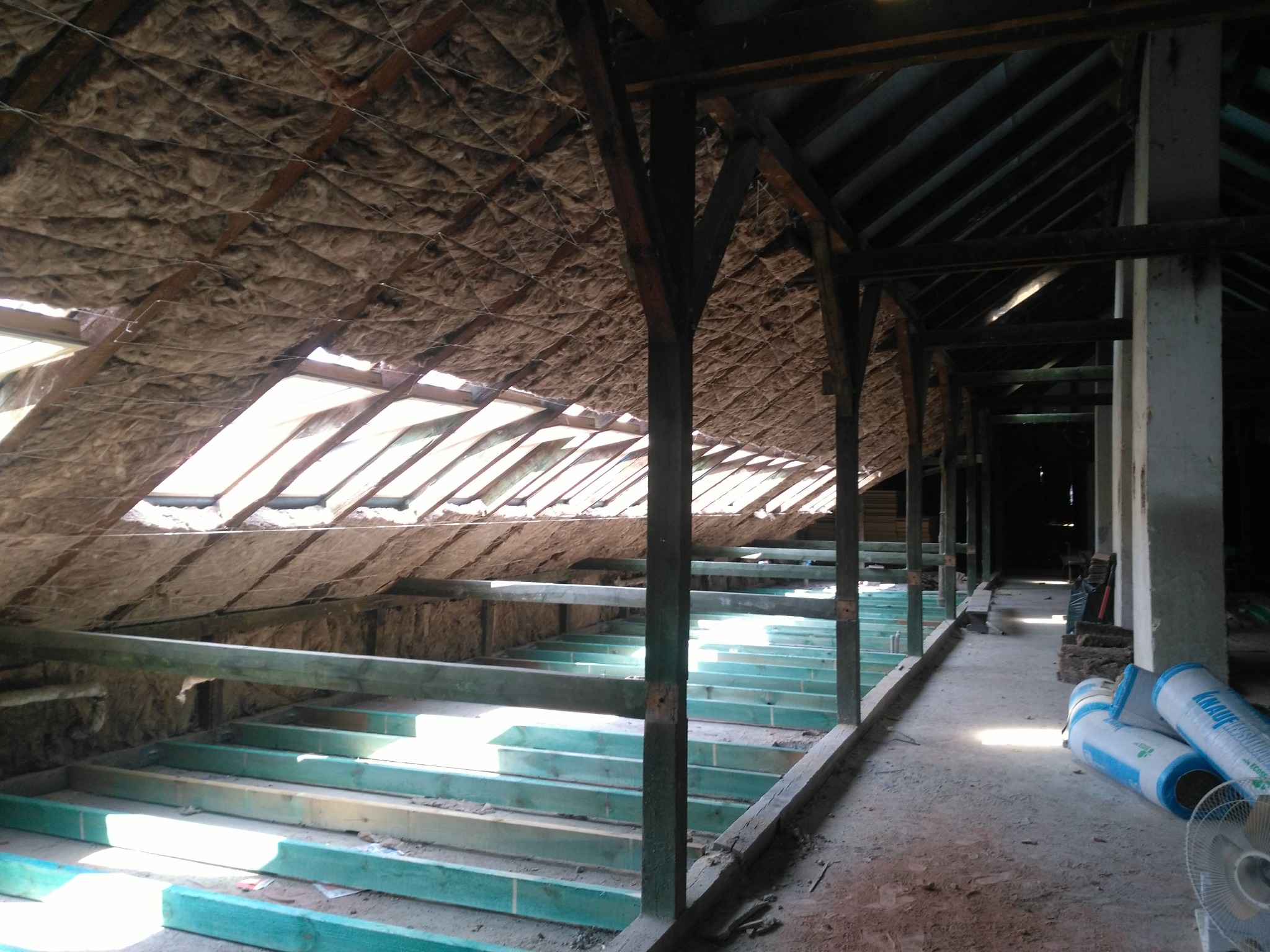 Poddasze w trakcie ocieplania wełną mineralną, widoczne drewniane krokwie i niebieskie rolki materiału izolacyjnego Knauf.