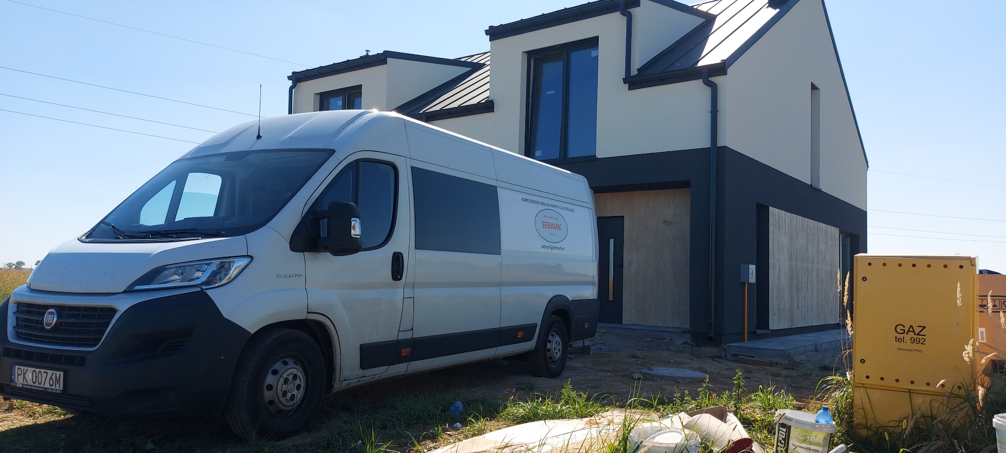 Nowoczesny dom w trakcie wykończenia elewacji, z zaparkowanym obok samochodem dostawczym firmy budowlanej z widocznym logo i danymi kontaktowymi na boku pojazdu.