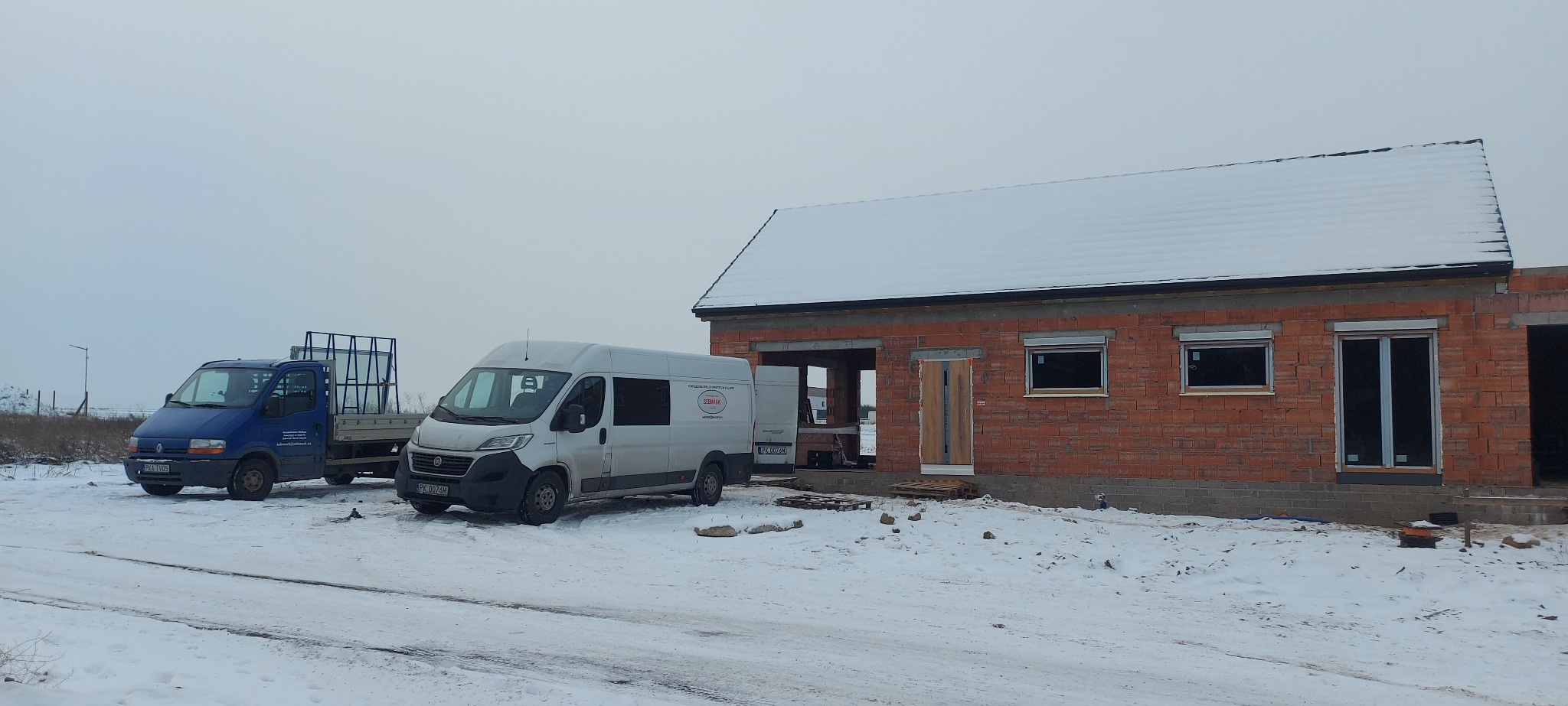 W zimowej scenerii w Kaliszu, budynek w trakcie budowy z zamontowanymi oknami z roletami zewnętrznymi oraz częściowo zamontowanymi drzwiami wejściowymi. Na pierwszym planie zaparkowane busy dostawcze.