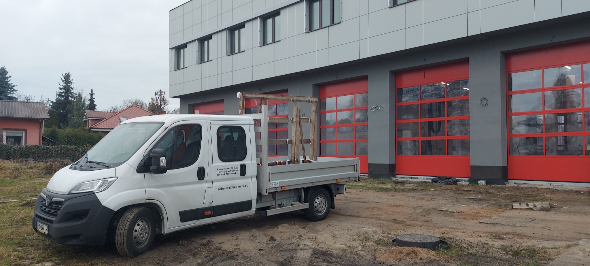 Biały samochód dostawczy z drewnianą konstrukcją na pace, zaparkowany przed budynkiem z szarymi ścianami i czerwonymi bramami garażowymi z oknami.