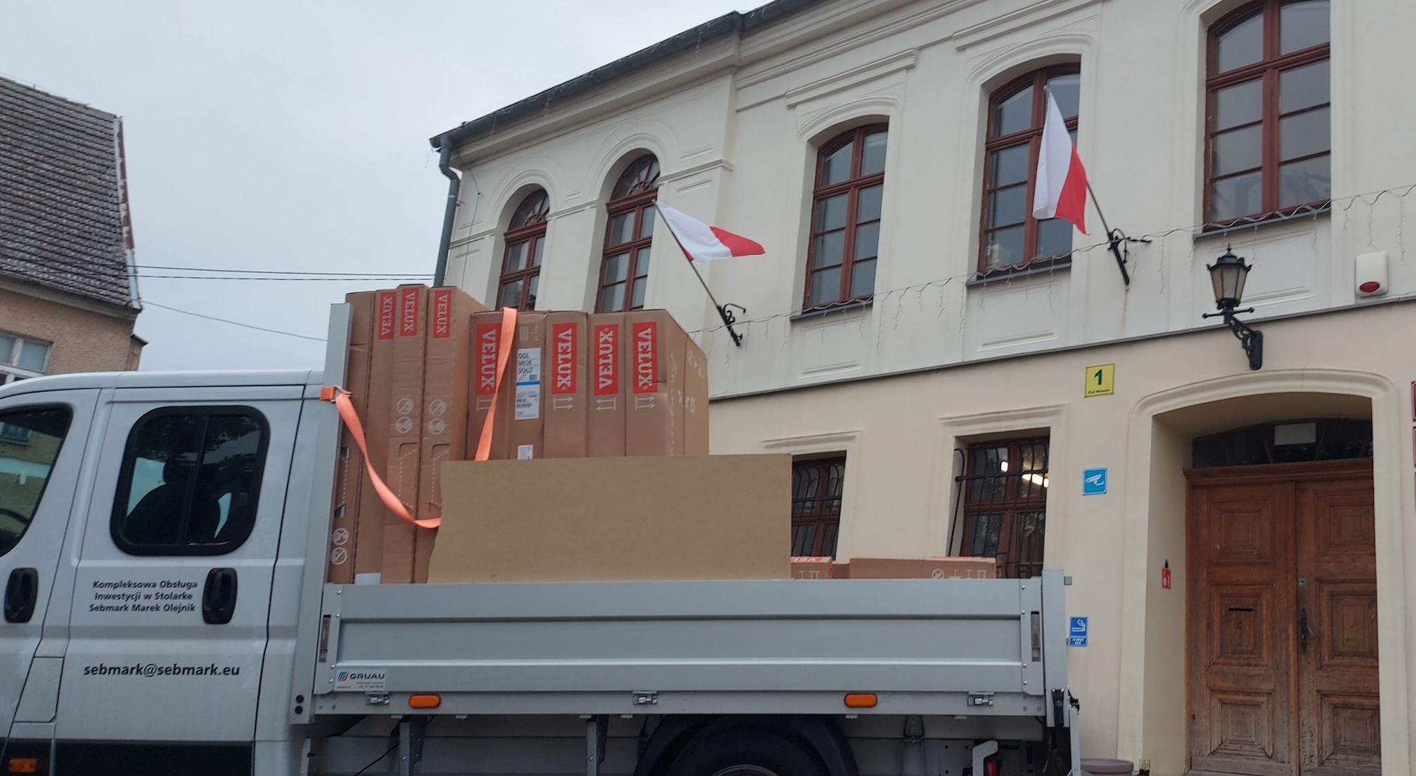 Dostawa okien dachowych Velux na pace samochodu dostawczego firmy Sebmark, na tle budynku z polską flagą.