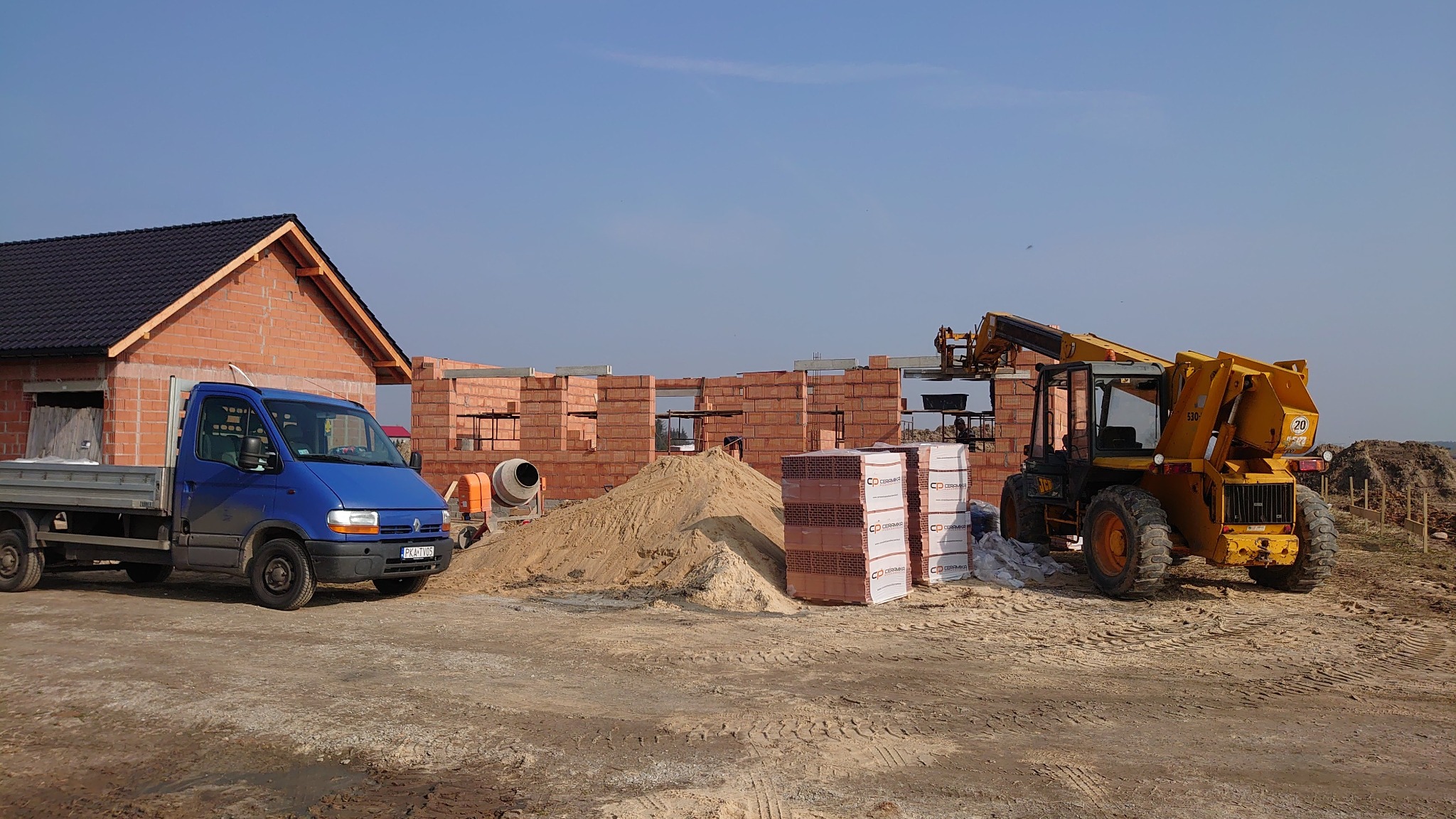 Wznoszenie murów domu z czerwonej cegły, widoczny niebieski samochód dostawczy, betoniarka i podnośnik teleskopowy JCB na terenie budowy w słoneczny dzień.