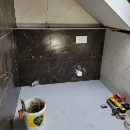 Łazienka w trakcie układania płytek, widoczne brązowe i beżowe płytki imitujące marmur, poziomica, wiadro z zaprawą klejową i system poziomowania płytek.