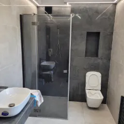 Nowoczesna łazienka z kabiną prysznicową typu walk-in, umywalką nablatową i podświetleniem LED podsufitowym, wykończona płytkami w odcieniach szarości i bieli.