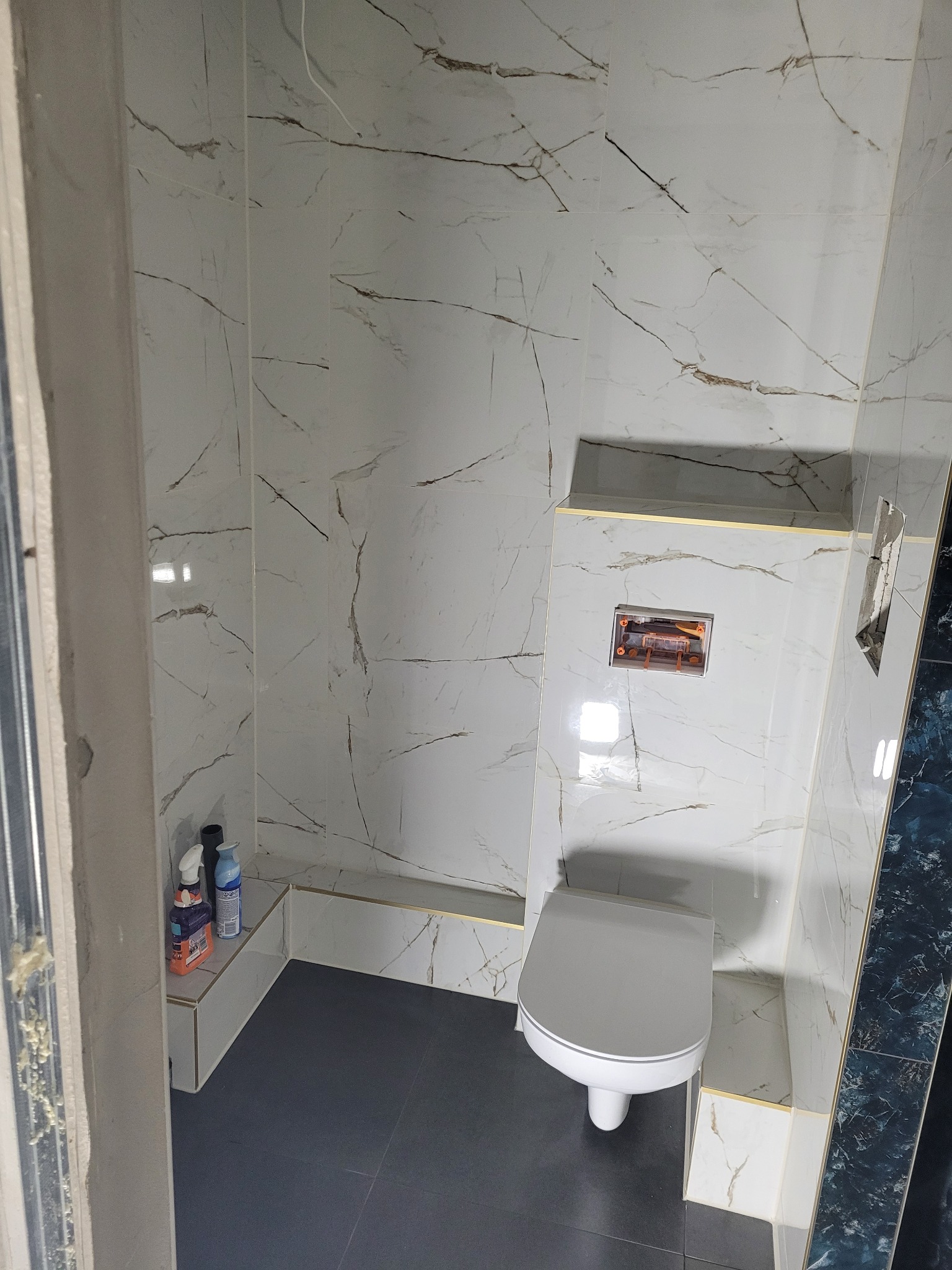 Wykończona toaleta w łazience z białymi płytkami imitującymi marmur, szarą podłogą i zabudową geberit z częściowo widoczną instalacją, widoczne środki czystości na półce.