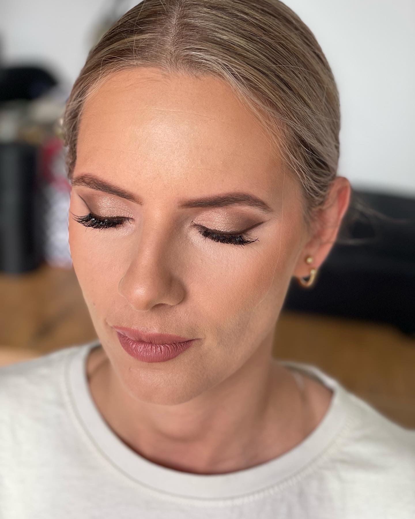 Portret kobiety z delikatnym, naturalnym makijażem oczu w odcieniach brązu, podkreślonymi sztucznymi rzęsami i ustami w kolorze nude, włosy upięte do góry, widoczny złoty kolczyk.