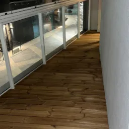 Zadaszony balkon z drewnianą podłogą i przeszkloną balustradą, widok na oświetlony chodnik z kostki brukowej na zewnątrz.