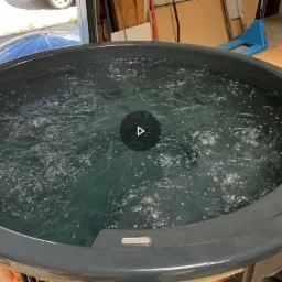 Szara balia ogrodowa wypełniona wodą, w tle widoczne elementy drewniane i narzędzia, sugerujące etap testowania przed wydaniem klientowi.