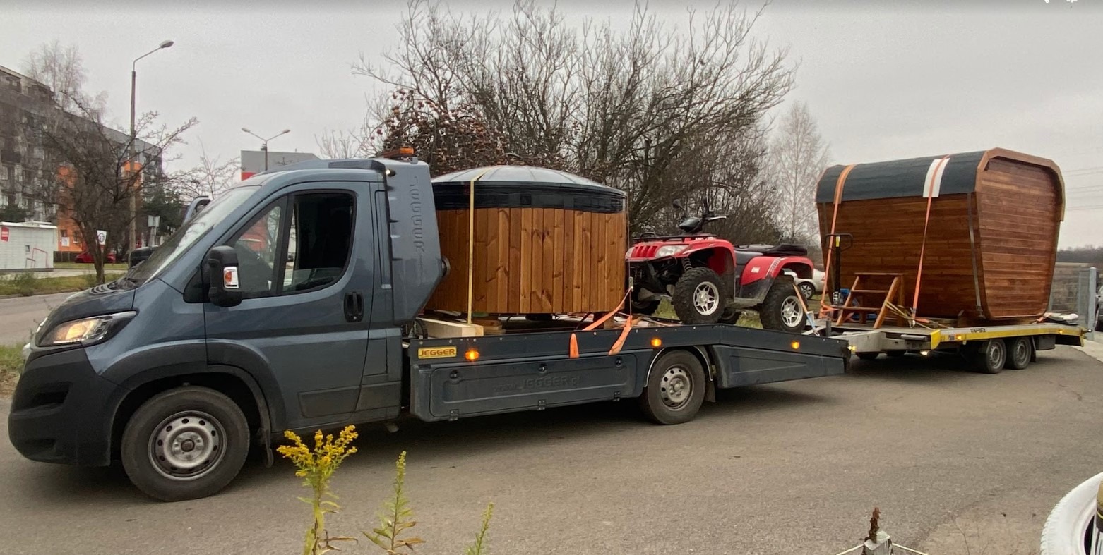 Transport sauny beczki i balii ogrodowej na lawecie z quadem zabezpieczone pasami transportowymi, widok z boku na szary samochód dostawczy na tle jesiennego krajobrazu.