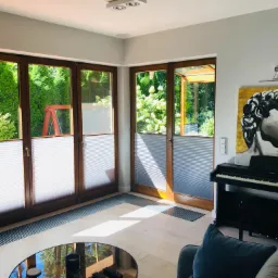 Jasne wnętrze z drewnianymi oknami i złożonymi tekstylnymi przesłonami okiennymi, widok na zielony ogród, na ścianie obraz z motywem twarzy i pianino.