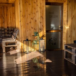 Mega Moc Paweł Drzeżdzon - Drewniane wnętrze sauny z ławami i stolikami z palet, dekoracyjne lampiony i roślina na stoliku, drzwi z napisem 'Wellness' i sylwetką osoby w saunie.