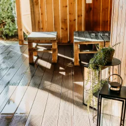 Drewniana konstrukcja z siedziskami z palet i dekoracyjną latarnią oraz rośliną w doniczce, podłoga z desek kompozytowych, widoczne refleksy światła na podłodze.