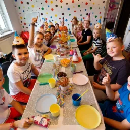 Grupa uśmiechniętych dzieci w kolorowych czapeczkach siedzi przy stole zastawionym przekąskami i napojami, świętując urodziny w pokoju z kolorowymi kropkami na ścianie.