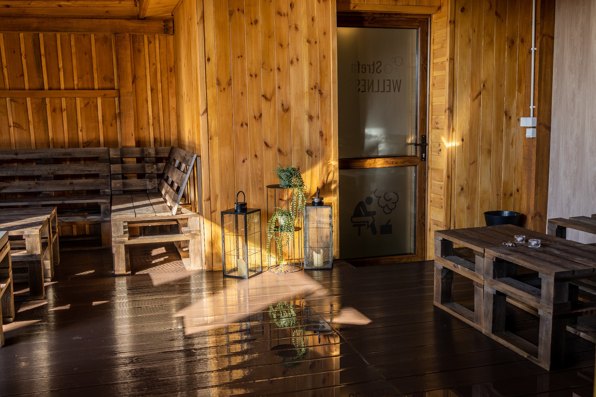 Drewniane wnętrze sauny z ławami i stolikami z palet, dekoracyjne lampiony i roślina na stoliku, drzwi z napisem 'Wellness' i sylwetką osoby w saunie.