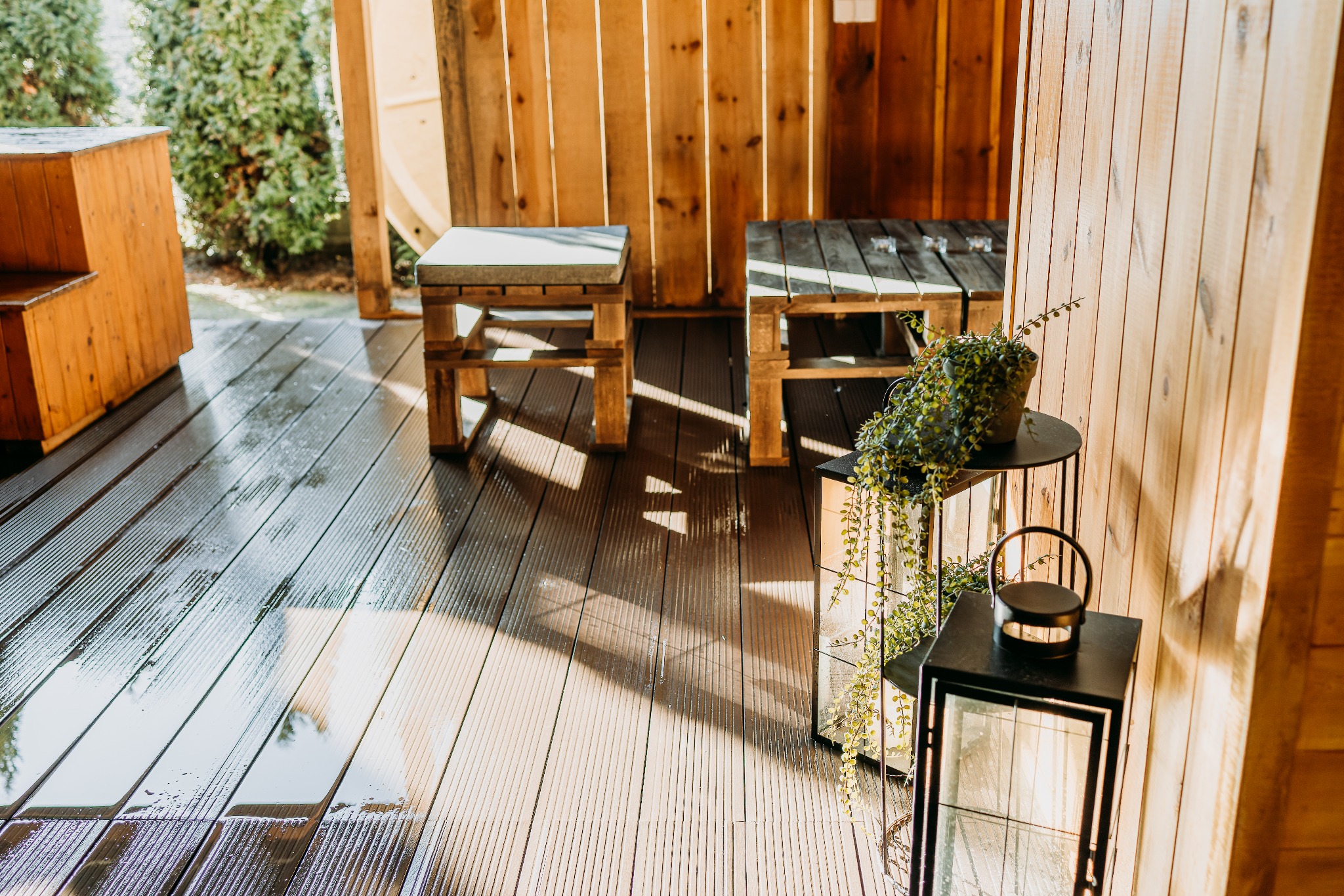 Drewniana konstrukcja z siedziskami z palet i dekoracyjną latarnią oraz rośliną w doniczce, podłoga z desek kompozytowych, widoczne refleksy światła na podłodze.