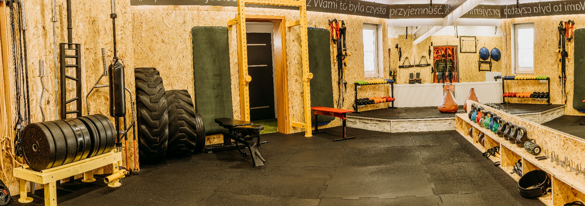 Przestronna sala do ćwiczeń z profesjonalnym sprzętem: ciężary, opony, kettlebells, worki bokserskie i drabinki gimnastyczne, wykończona płytami OSB i gumową podłogą.