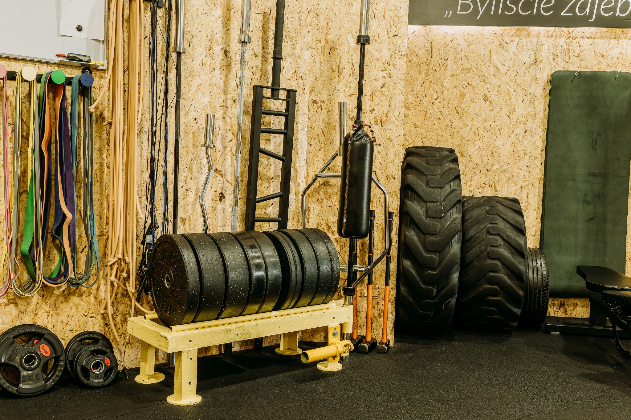 Wyposażenie siłowni: obciążenia na stojaku, opony, worki treningowe, liny oporowe zawieszone na ścianie z płyty OSB