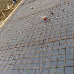 Zbrojenie fundamentu z siatki stalowej na folii, widoczne wystające elementy instalacji i przygotowany szalunek w tle.