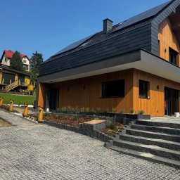 S-Bud Firma Ogólnobudowlana Skibiński Krzysztof - Nowoczesny dom jednorodzinny z drewnianą elewacją i ciemnym dachem w Turzy Śląskiej, widok z kamiennego podjazdu.