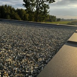 S-Bud Firma Ogólnobudowlana Skibiński Krzysztof - Dach pokryty drobnym żwirem w słoneczny dzień, widoczne metalowe obramowanie dachu, drzewa i pole w tle.