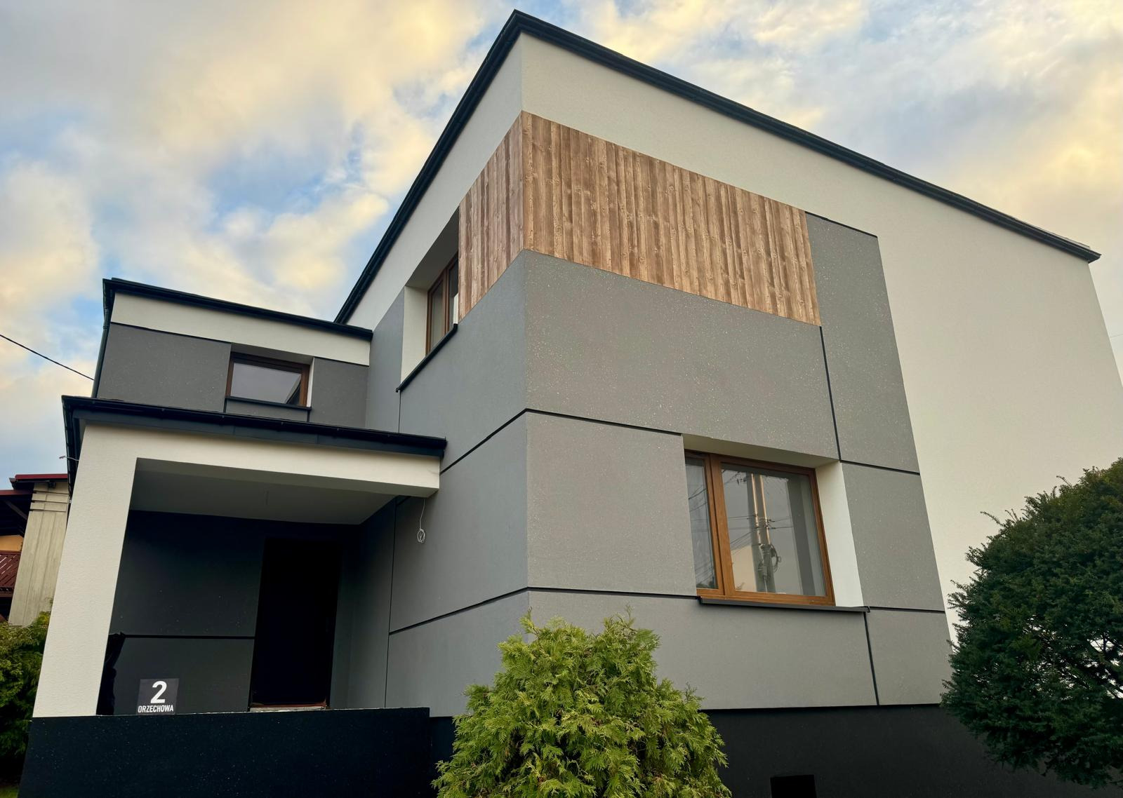 Nowoczesny dom jednorodzinny z elementami dekoracyjnymi z drewna na elewacji, antracytowe obramowania okien i dachu, oraz numer posesji '2 Orzechowa' przy wejściu.