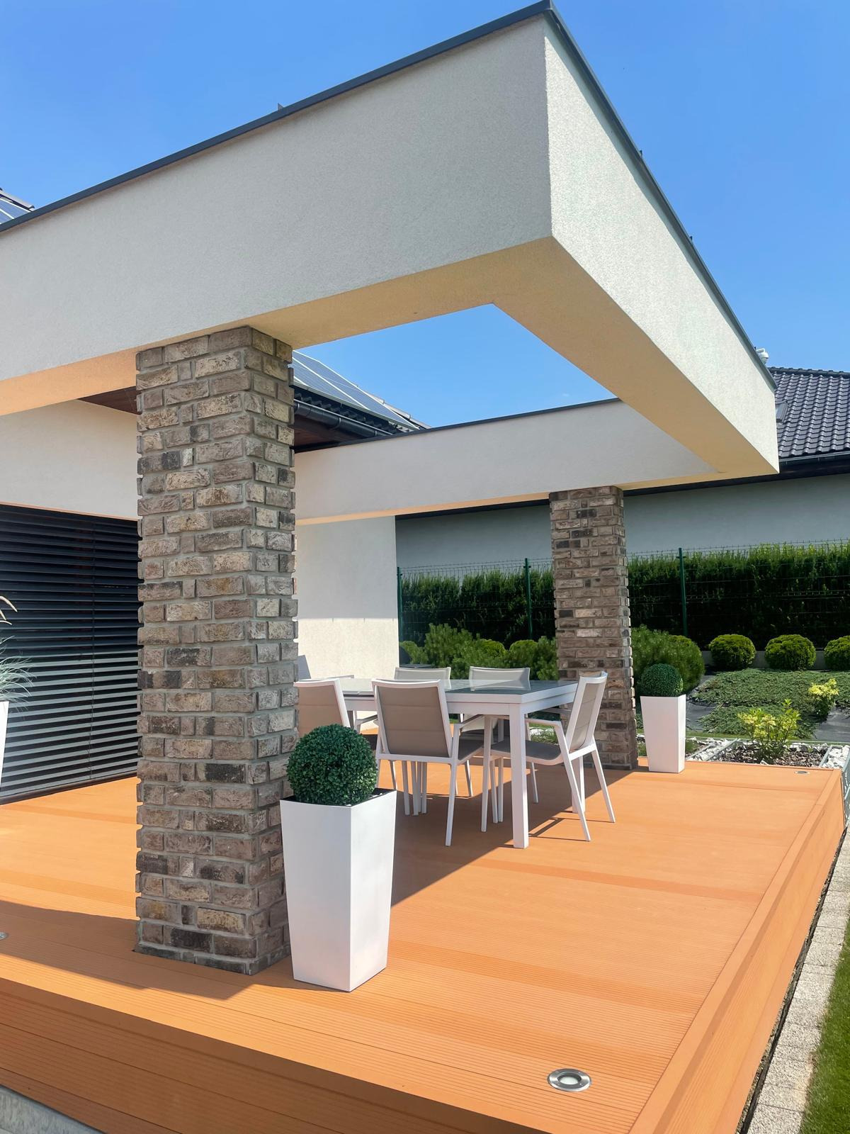 Nowoczesna, zadaszona przestrzeń tarasowa z kolumnami z cegły klinkierowej, drewnianą podłogą i białymi donicami z kulistymi roślinami, uzupełniona białym zestawem mebli ogrodowych i starannie...