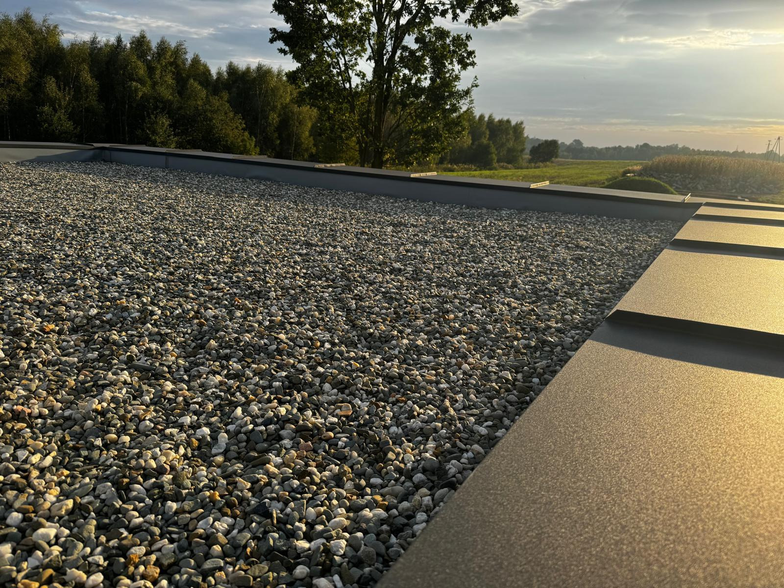 Dach pokryty drobnym żwirem w słoneczny dzień, widoczne metalowe obramowanie dachu, drzewa i pole w tle.