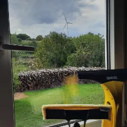 Żółty odkurzacz do okien Kärcher na parapecie, widok na wiejski krajobraz z stosem drewna i turbiną wiatrową za oknem, szyba w trakcie czyszczenia.