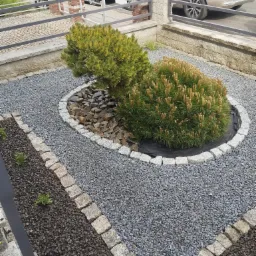 Ogródek z dwoma karłowatymi sosnami otoczonymi kamieniami, wysypany szarym żwirem, z obrzeżem z kostki granitowej, w tle zaparkowane samochody.