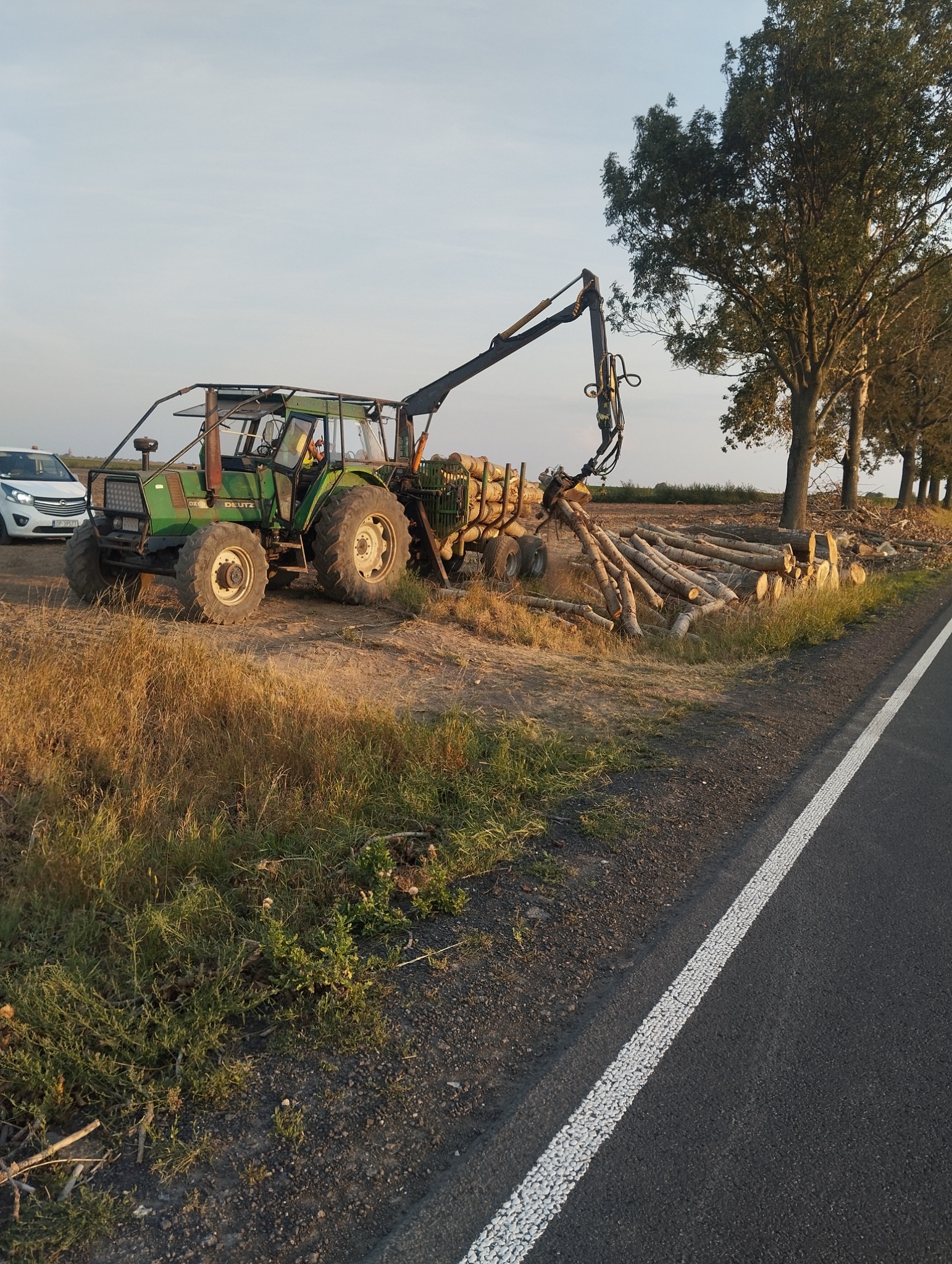 Zielony traktor Deutz z hydraulicznym ramieniem załadowuje ścięte pnie drzew na przyczepę obok asfaltowej drogi z białą linią, w tle rząd drzew i biały samochód.