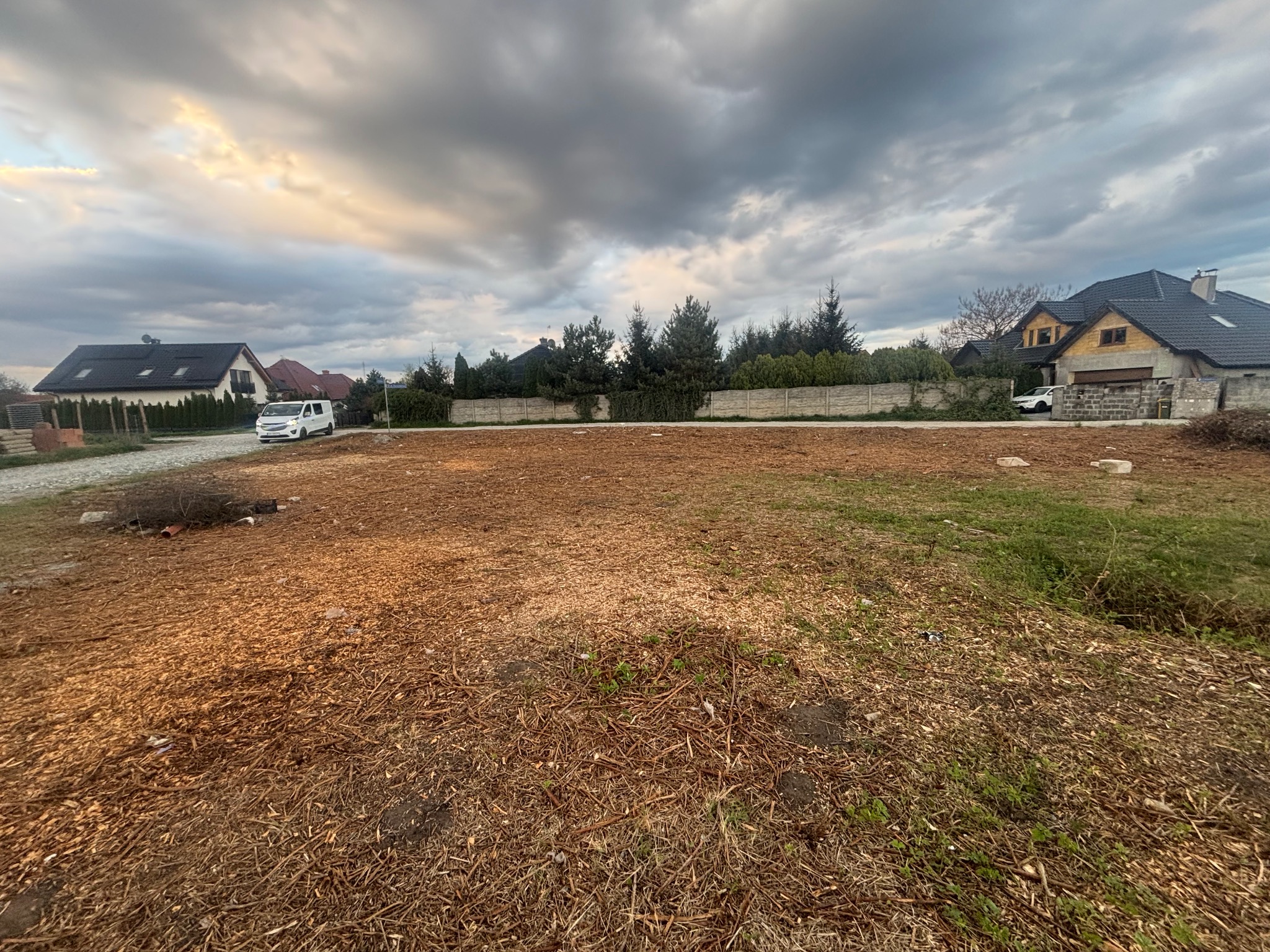 Działka po wycince drzew, pokryta zrębkami, z widocznymi zabudowaniami w tle pod pochmurnym niebem.