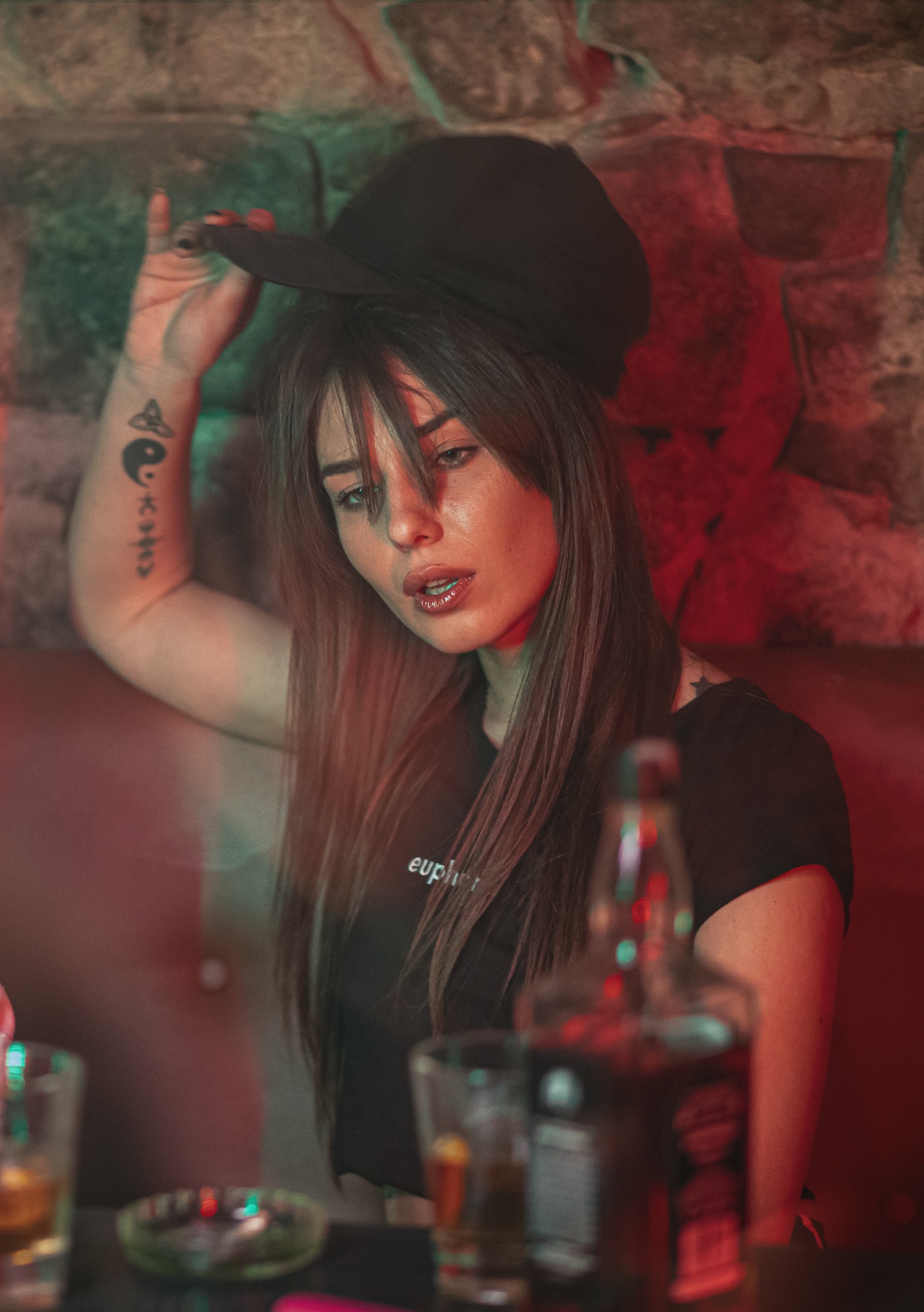 Portret kobiety z tatuażem na ramieniu, trzymającej czarną czapkę z daszkiem w barze, butelka Jacka Danielsa na stole, oświetlenie w odcieniach czerwieni i zieleni.