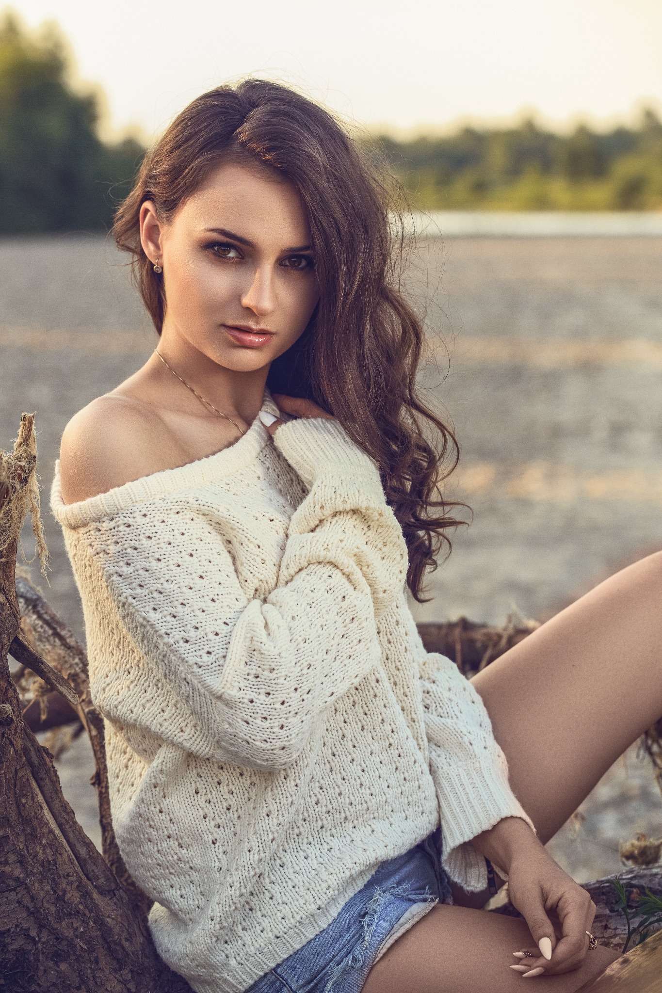 Portret młodej kobiety w kremowym swetrze z odkrytym ramieniem, siedzącej na pniu drzewa na tle rozmytej zieleni i piasku.
