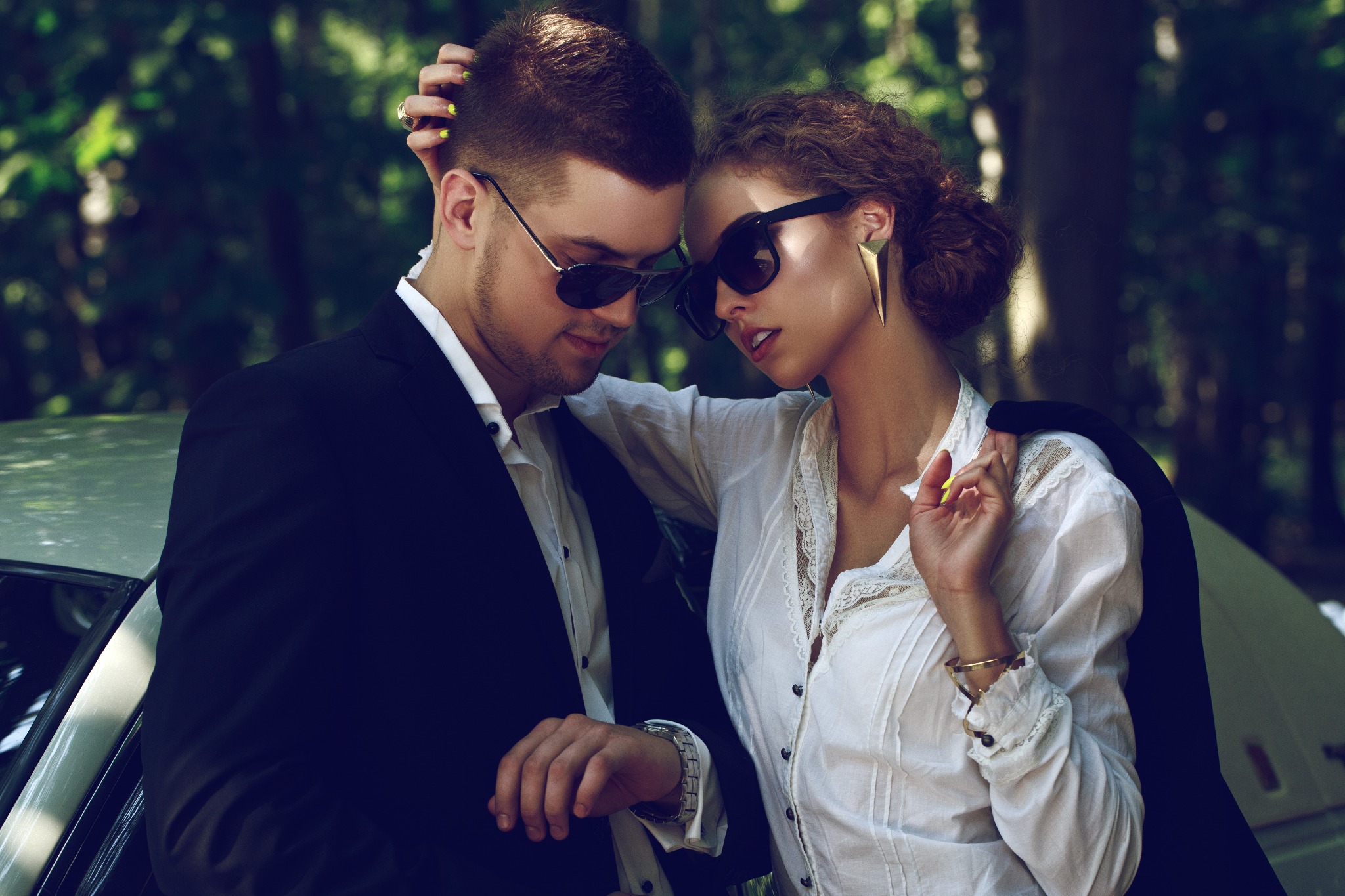 Elegancka para w okularach przeciwsłonecznych pozuje przy samochodzie: mężczyzna w garniturze sprawdza godzinę na zegarku, kobieta w białej bluzce z koronką i dużych kolczykach opiera się o niego.