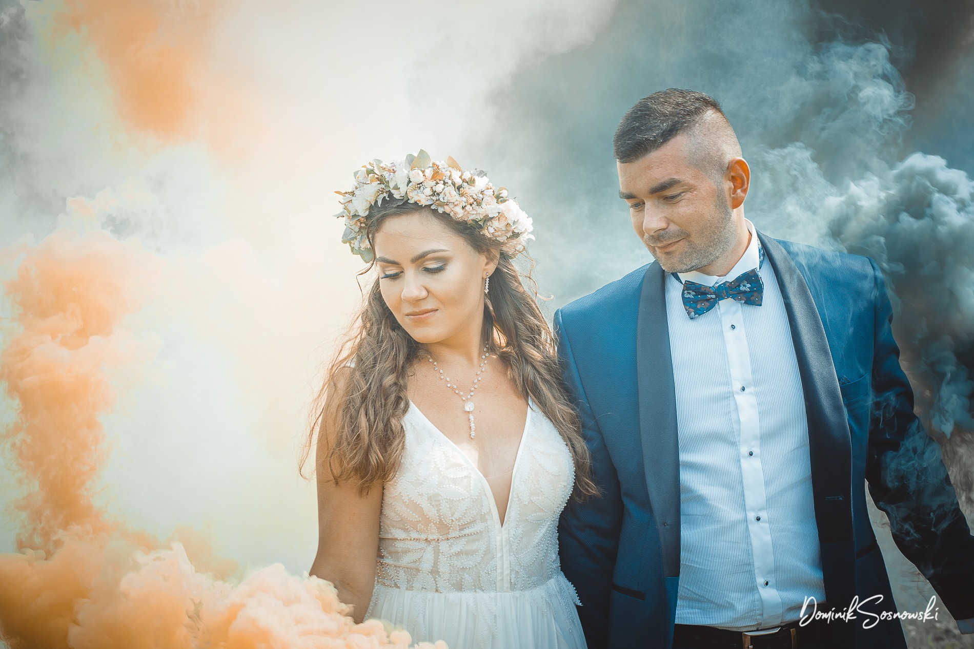 Para młoda w otoczeniu kolorowych dymów: panna młoda z wiankiem na głowie i naszyjnikiem, pan młody w granatowym garniturze z muchą.