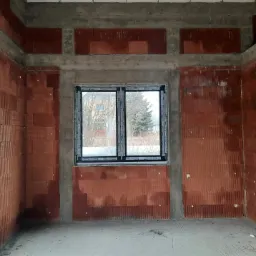 Surowy stan wnętrza z wstawionym oknem dwuszybowym z czarną ramą, widok na zewnątrz na drzewa w tle, ściany z czerwonej cegły i betonowe nadproża.