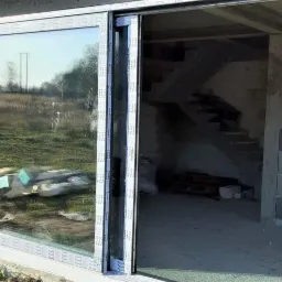 Wnętrze surowego budynku z zamontowanymi przesuwnymi drzwiami tarasowymi z widokiem na zewnątrz. Widoczne bloczki betonowe, schody w stanie surowym i elementy konstrukcyjne.