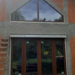 Montaż okna z brązowymi ramami i roletą zewnętrzną w surowym stanie budynku z cegły, widoczne rusztowanie drewniane z lewej strony.