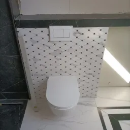 Biała toaleta wisząca z podwójnym przyciskiem spłukującym na ścianie wyłożonej białymi płytkami z ciemnymi akcentami, widoczne fragmenty podłogi i ściany z ciemnego marmuru.