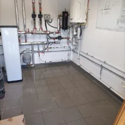 Widok na kotłownię z zainstalowanym piecem gazowym, zasobnikiem wody, naczyniem wzbiorczym i rozbudowaną instalacją hydrauliczną z rur miedzianych i ocynkowanych, schemat kotłowni na ścianie.