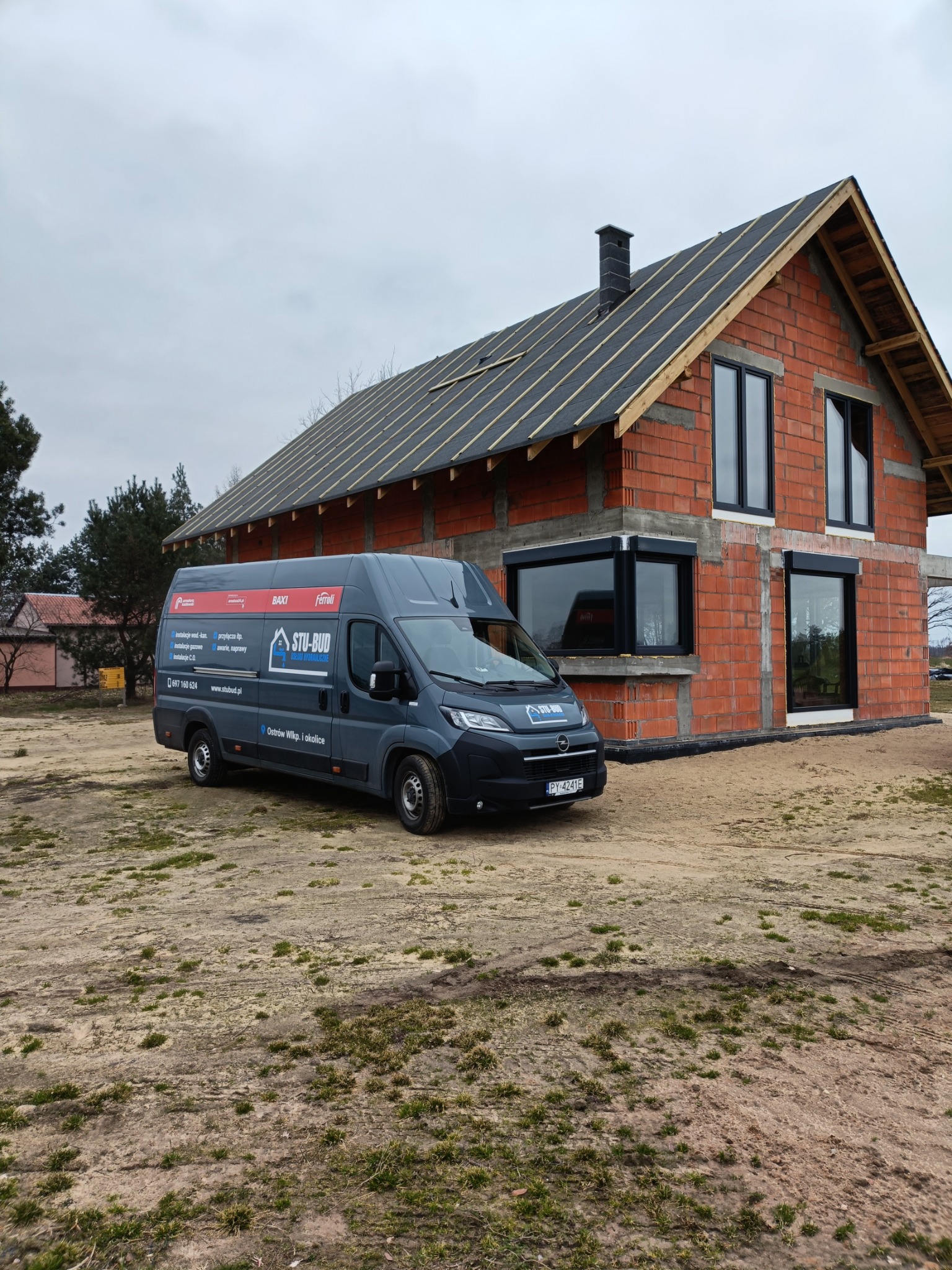 Szary bus firmy instalacyjnej STU-BUD na tle budowanego domu z czerwonej cegły z zamontowanymi czarnymi oknami i przygotowanym dachem pod pokrycie.